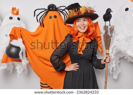 Horizontal shot of cheerful witch with orange hair wears black dress poses around creepy creatures holds broom with crow prepares for Halloween party enjoys celebration. Trick or treat conept