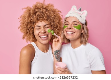 Horizontal shot of cheerful women friends undergo beauty procedures apply hydrogel patches and facial mask for rejuvenation and skin treatment dressed casually isolated over pink background. - Powered by Shutterstock