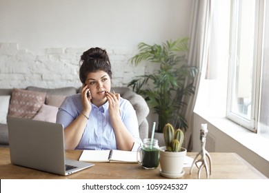 Horizontal shot of concentrated chubby young woman with hair bun multitasking at home office, sitting at desk in front of open laptop computer, making notes in diary and having phone conversation