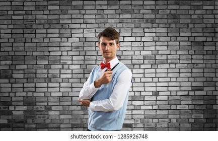 Horizontal shot of confident and young businessman in smart-casual wear smoking pipe while standing against gray brick wall on background.