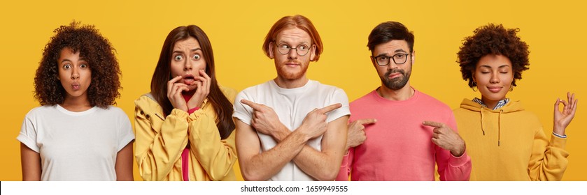 Horizontal shot of confused man points sideways, bearded male indicates at himself, two Afro American women and scared European lady. All people stand in one row, isolated over yellow background