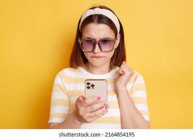 Horizontal shot of confused young woman wearing striped t-shirt standing with mobile phone and looking at display with puzzlement, posing isolated over yellow background.