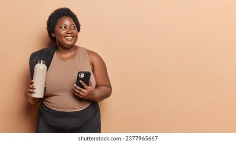 Horizontal shot of curly African American plus size woman holding sports bottle with water and smartphone satisfied after cardio training dressed in activewear isolated over brown background - Powered by Shutterstock