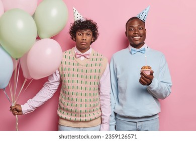 Horizontal shot of curly haired Hindu man and black man dressed in festive clothing holds cupcake with candle holds bunch of colorful balloons celebrates special occasion isolated over pink background