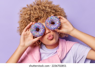 Horizontal shot of curly haired woman covers eyes with delicious glazed doughnuts keeps lips rounded poses with sweet delicious dessert poses against purple background. Unhealthy eating concept