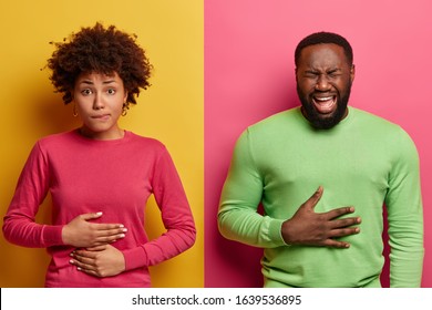 Horizontal shot of dark skinned couple touch stomachs, suffer from chronic gastritis, being hungry has displeased face expressions, isolated over yellow and pink background. Healthcare concept