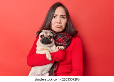 Horizontal shot of disappoited brunette Asian woman looks frustrated holds pug dog sad because of bad weather cannot go for walk dressed in casuall clothes isolated over vivid red background.