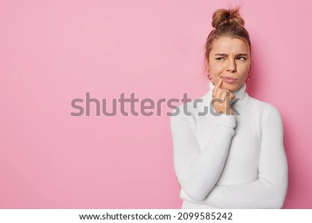 Horizontal shot of discontent sullen young woman looks thoughtfully away purses lips wears casual white turtleneck isolated over pink background with copy space for your promotional content.