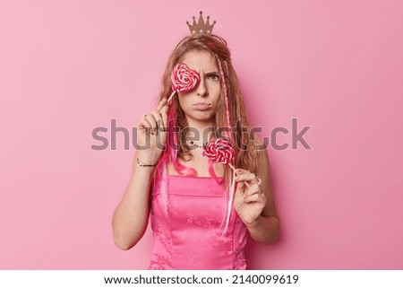 Horizontal shot of discontent young woman with long hair wears small crown and dress holds heart lollipops has sulking expression isolated over pink background. Angry princess with caramel candies