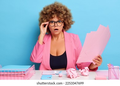 Horizontal shot of displeased business woman in modern pink blazer and eyeglasses holds bunch of papers, makes financial report looks grumpy, sits at messy workspace, isolated over blue wall. eople