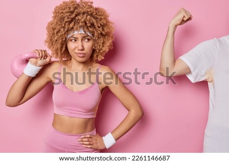 Horizontal shot of displeased woman with curly hair lifts weight dressed in sportswear smirks face smell umplesant stinch spends free time in gym isolated on pink background. Active lifestyle concept
