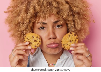 Horizontal Shot Of Displeased Woman Has Sulking Upset Expression Holds Two Cookies With Chocolate Feels Temptation To Eat Delicious Appetizing Snack Poses Indoor. Harmful Food And Bad Mood Concept