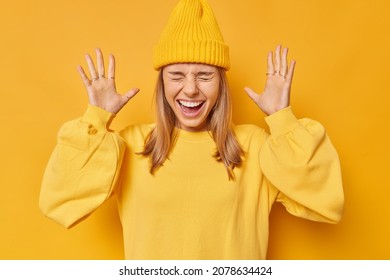 Horizontal shot of emotional millennial girl exclaims loudly keeps palms raised reacts on awesome news wears hat and jumper isolated over yellow background. Human emotions and reactions concept - Shutterstock ID 2078634424