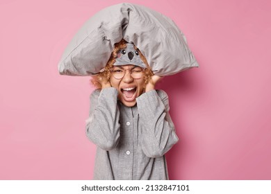 Horizontal Shot Of Emotional Woman Exclaims Loudly Cannot Fall Asleep Because Of Noise Keeps Soft Pillow Over Head Covers Ears Dressed In Nightwear Isolated Over Pink Background. Sleep Interruption