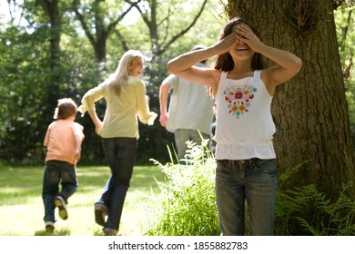 Horizontal shot of a family of four playing hide and seek in the forest with the girl covering eyes while her family runs away.