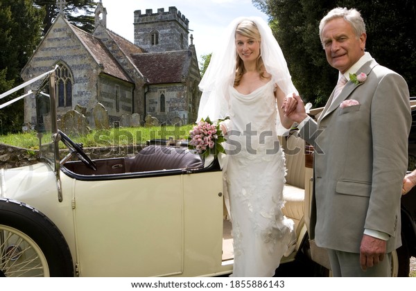 Horizontal shot of a\
father helping the bride out of a vintage car by church smile at\
the camera with copy\
space.