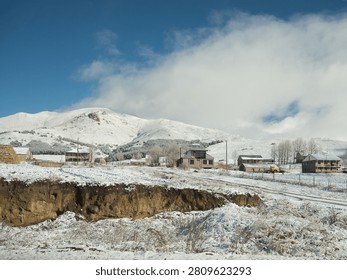 A horizontal shot featuring an eroded dark soil bank in the foreground with a snow-covered village and majestic white mountains of Armenia behind.