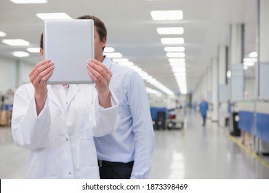 Horizontal Shot Of A Female Scientist Holding Up A Digital Tablet To Face As A Businessman Besides Looks On With Obscured Faces On A Factory Floor.