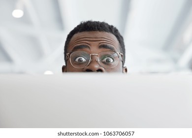 Horizontal Shot Of Funny Dark Skinned Male In Big Round Glasses, Looks From Laptop Computer, Has Funny Expression, Shocked To Have Deadline And Much Work. People, Facial Expressions, Technology