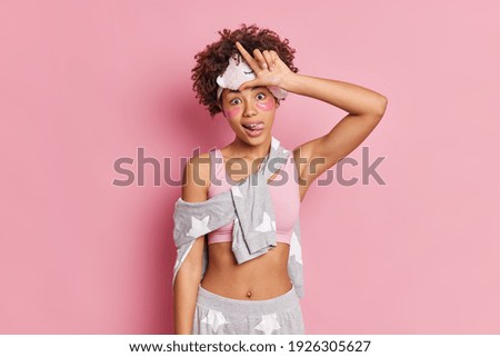 Horizontal shot of funny dark skinned African American woman makes loser gesture sticks out tongue teases someone dressed in sleepwear wears beauty pads isolated over pink background. Body language