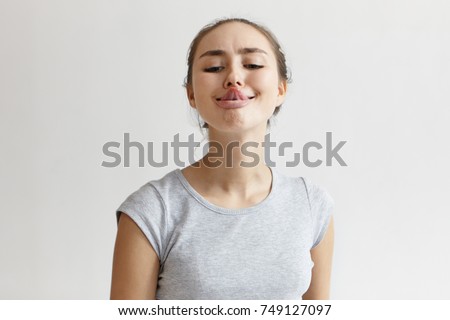 Horizontal shot of funny humorous playful young European woman in grey t-shirt sticking out her tongue, trying to touch nose with its tip. People, youth, leisure, joy, fun and entertainment concept.