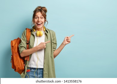 Horizontal Shot Of Glad Caucasian Woman Points Aside With Both Fore Fingers, Advertises Something Aside, Dresed In Stylish Clothes, Carries Rucksack, Listens Music In Modern Headphones. Promotion