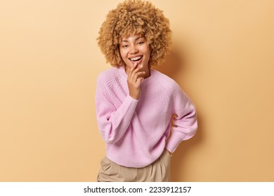 Horizontal shot of good looking glad woman looks with curious expression keeps hand on chin smiles toothily wears casual pink jumper and trousers keeps hand in pocket isolated over beige wall