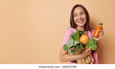 Horizontal shot of good looking woman with eastern appearance carries paper bag full of fresh vegetables and plastic bottle of smoothie looks happily away isolated over beige background blank space - Shutterstock ID 2203227339