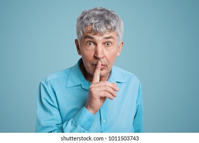 Horizontal shot of grey haired male looks with puzzled expression at camera, keeps fore finger on lips, asks to be quiet and mute, demands hush, dressed in elegant formal shirt isolated over blue wall