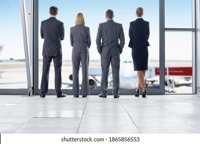 Horizontal Shot Of A Group Of Four Business People In Suits Standing In Front Of An Airport Window With Their Backs To The Camera