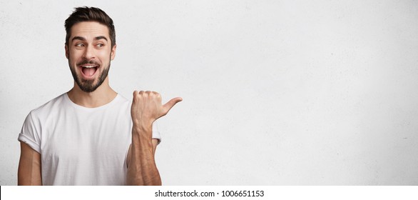 Horizontal shot of handsome bearded young brunet male model has appealing appearance, dressed casually, indicates with thumb aside, demostrates something excited and positive at blank copy space