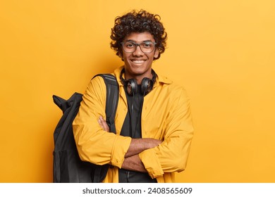 Horizontal shot of handsome curly haired Hindu man dreessed in casual clothing carries black rucksack on shoulder headphones around neck isolated over vivid yellow background. Studying concept - Powered by Shutterstock