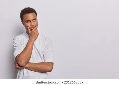 Horizontal shot of handsome dark skinned man ponders solution to problem keeps hand on chin stands in thoughtful pose wears casual t shirt isolated over white background copy space for your promotion - Shutterstock ID 2312668557