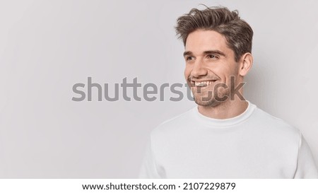 Horizontal shot of handsome pleased European man looks away with happy smile on face dressed casually feels satisfied notices something into distance isolated over white bacground blank copy space