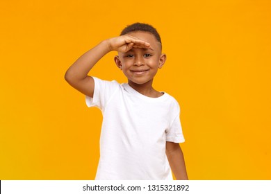 Horizontal shot of handsome positive eight year old dark skinned male child with skinny slim body posing at yellow studio wall, keeping palm on his forehead, looking into distance and smiling