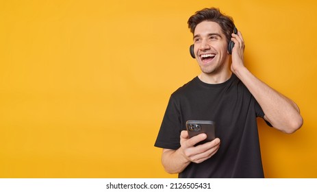 Horizontal shot of happy adult man dressed in casual black t shirt listens music via headphones enjoys favorite audio track isolated over yellow background blank copy space for your promotion - Shutterstock ID 2126505431