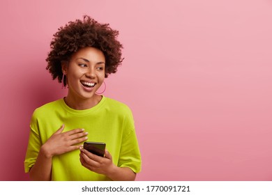 Horizontal Shot Of Happy African American Woman Uses Social Media On Smartphone, Looks Away With Broad Smile, Uses Free Wi Fi And Messages In Social Networks, Poses Over Rosy Wall, Blank Space Aside