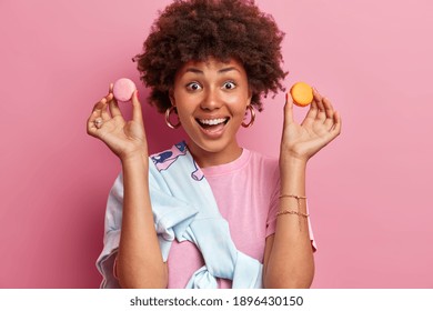 Horizontal shot of happy Afro American woman holds two macaroons giggles positively dressed in casual clothes has sweet tooth isolated over pink background eats junk food with much calories.