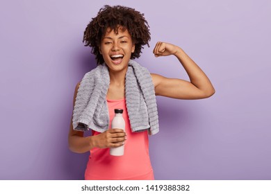 Horizontal shot of happy Afro woman shows biceps, satisfied after active training, holds bottle of water, enjoys healthy lifestyle, isolated over purple background. Sport and exercising concept