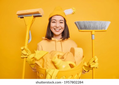 Horizontal Shot Of Happy Asian Girl Helps Mom To Do Housework Holds Mop And Broom Has Glad Expression Wears Hat Sweatshirt Protective Rubber Gloves Isolated Over Yellow Background. Domestic Work