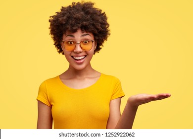 Horizontal shot of happy black woman with trendy shades, crisp hair, dressed in casual clothing, holds copy space, stands against yellow background. People, ethnicity and advertisement concept