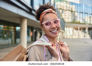 Horizontal shot of happy curly haired woman wears stylish pink sunglasses headband and jacket carries paper shopping bags glad to return from mall makes successful purchases. Consumerism concept