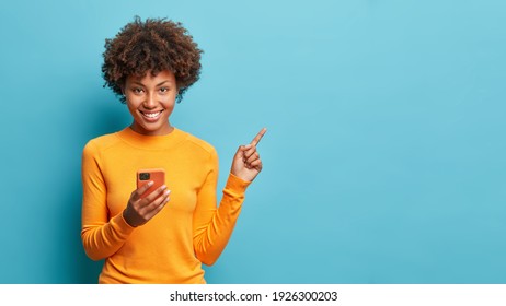 Horizontal shot of happy dark skinned Afro American woman enjoys mobile communication and modern technologies poses against blue background points away on free space for your advertising content