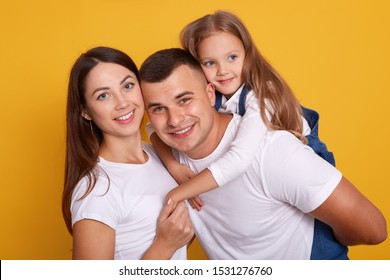 Horizontal shot of happy family wearing white shirts, stand smiling isoalted over yellow studio background, father piggybacking her adorable female kid. Relationship, happyness and devotion concept. - Shutterstock ID 1531276760