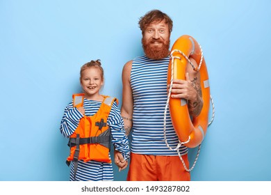 Horizontal Shot Of Happy Father And Child In Protective Lifejacket, Carry Lifering, Spend Summer Vacation Together, Learn Swimming, Express Good Emotions, Isolated Over Blue Background. Fatherhood