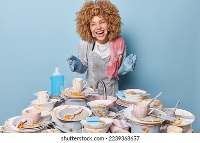 Horizontal shot of happy housewife wears apron and rubber gloves stands near pile of dirty dishes has soap bubbles on face looks gladfully aside isolated over blue background. Housework concept - Shutterstock ID 2239368657