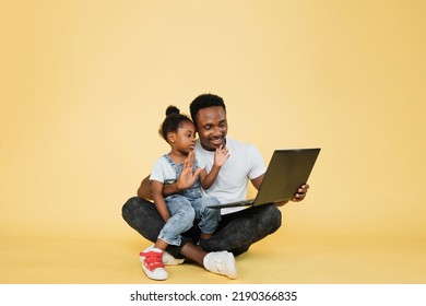 Horizontal shot of happy joyful African family, young father with cute little kid daughter, sitting on the floor and waving hands while watching a video lesson on laptop pc on yellow studio - Powered by Shutterstock