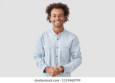 Horizontal shot of happy mixed race male with shining smile, feels glad as recieves bonus and praise for good work, has specific appearance, Afro hairstyle, dressed in fashionable white shirt