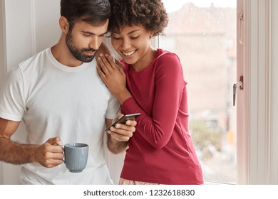 Horizontal Shot Of Happy Mixed Race Family Couple Enjoy Domestic Atmosphere, Drink Coffee, Read News In Internet Via Cell Phone, Stand Against Window Background. Diverse Woman And Man With Device