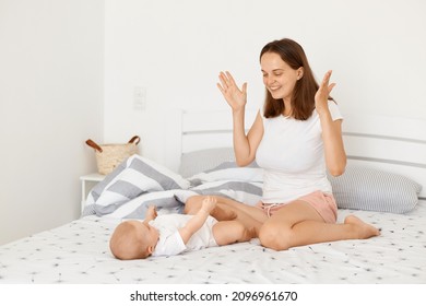 Horizontal shot of happy playful mother wearing white casual t shirt sitting on bed in bedroom with toddler baby, playing hide-and-seek with her little child.